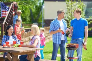 People at an outdoor BBQ. Two women sitting at picnic table talking. Two men standing by the BBQ talking and kids in the background
