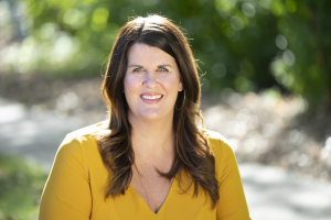 - Our Lady of Peace A headshot of Lindsey wearing a mustard colored top sitting in a garden.