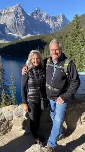 A couple standing in front of a lake with rolling green hills and then mountains in the distance. Both are smiling