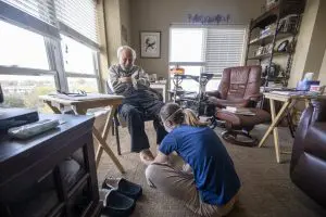 older man with white hair and beard sitting in a chair in his home. He is wearing a plaid shirt and dark pants with no socks or shoes. There is a women with a ponytail wearing a blue t-shirt and grey pants checking his feet for sores and providing a massage.