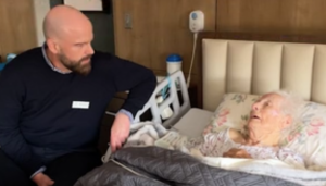 a bald man with a beard is leaning against a hospital bed talking to a frail older lady laying in bed