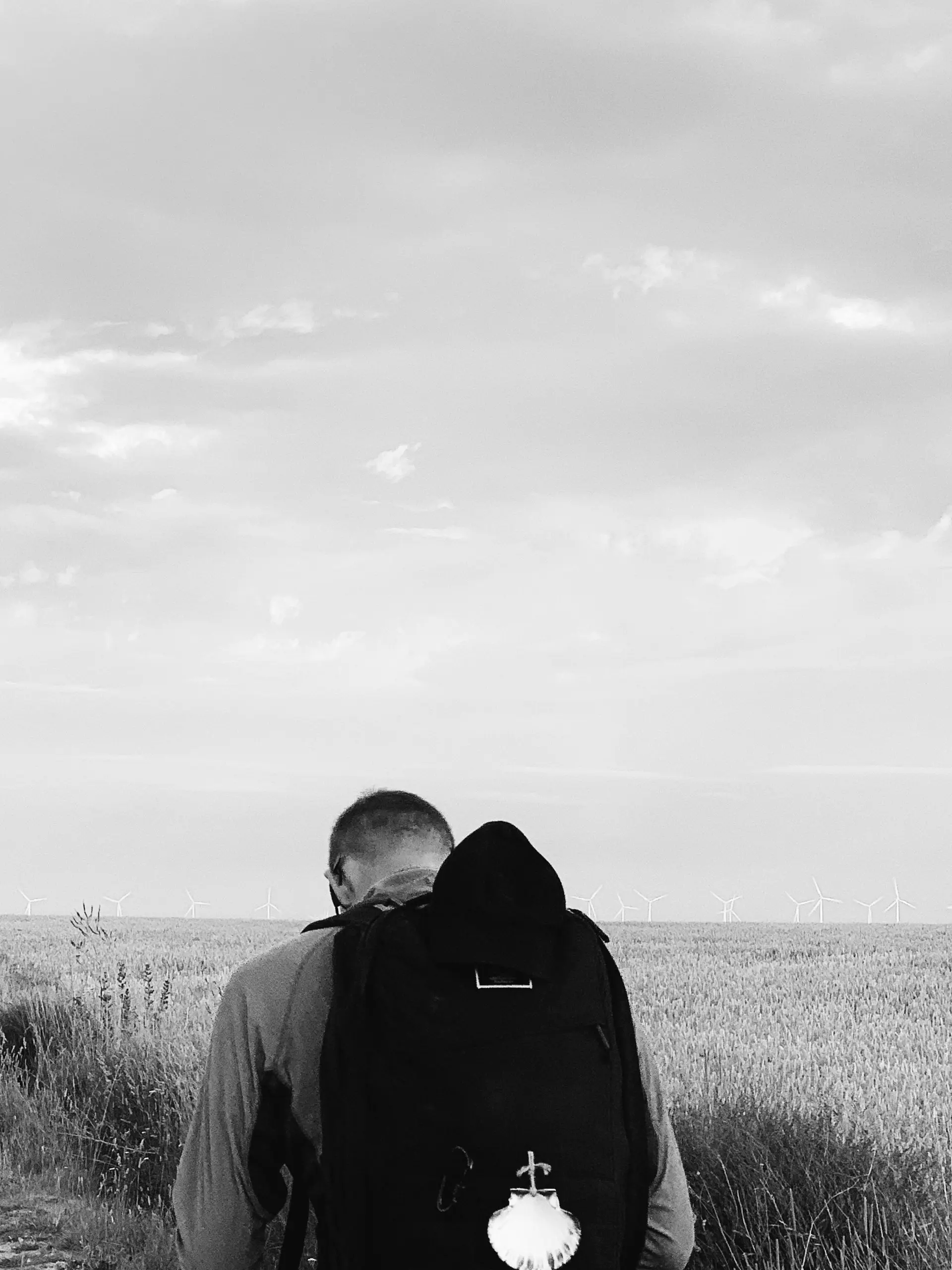 black and white photo the back of a man, head down who is wearing a black backpack standing in front of a field