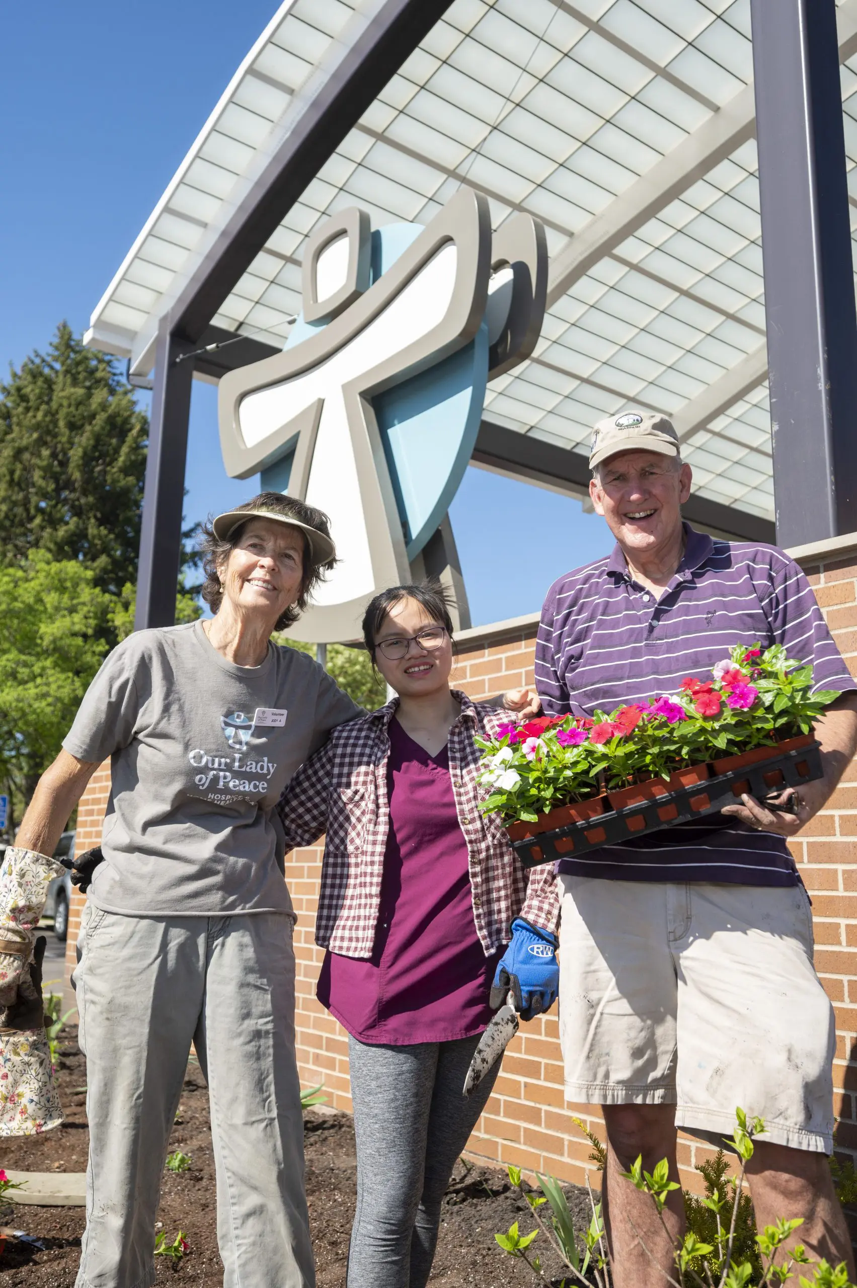2 woman and a man holding flowers standing in front of the our lady of peace logo