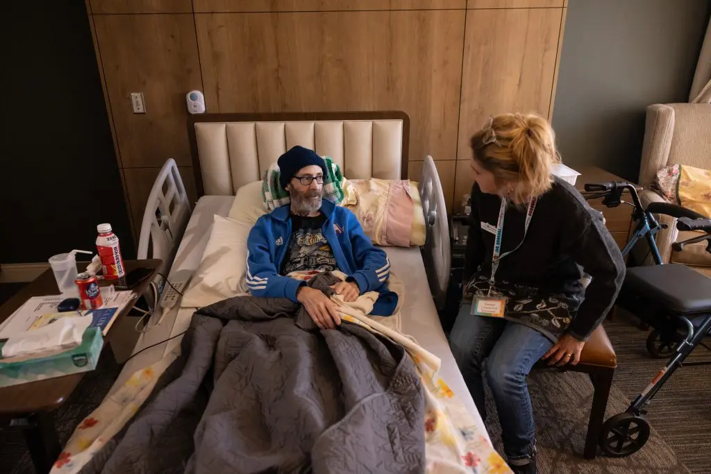 Volunteer_Patient - Our Lady of Peace man laying in a bed with a blue sweatshirt on, stocking cap and is talking to a woman with a blond ponytail
