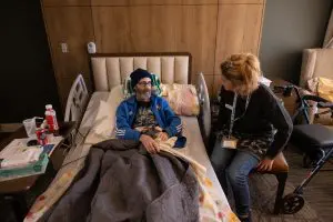 man laying in a bed with a blue sweatshirt on, stocking cap and is talking to a woman with a blond ponytail