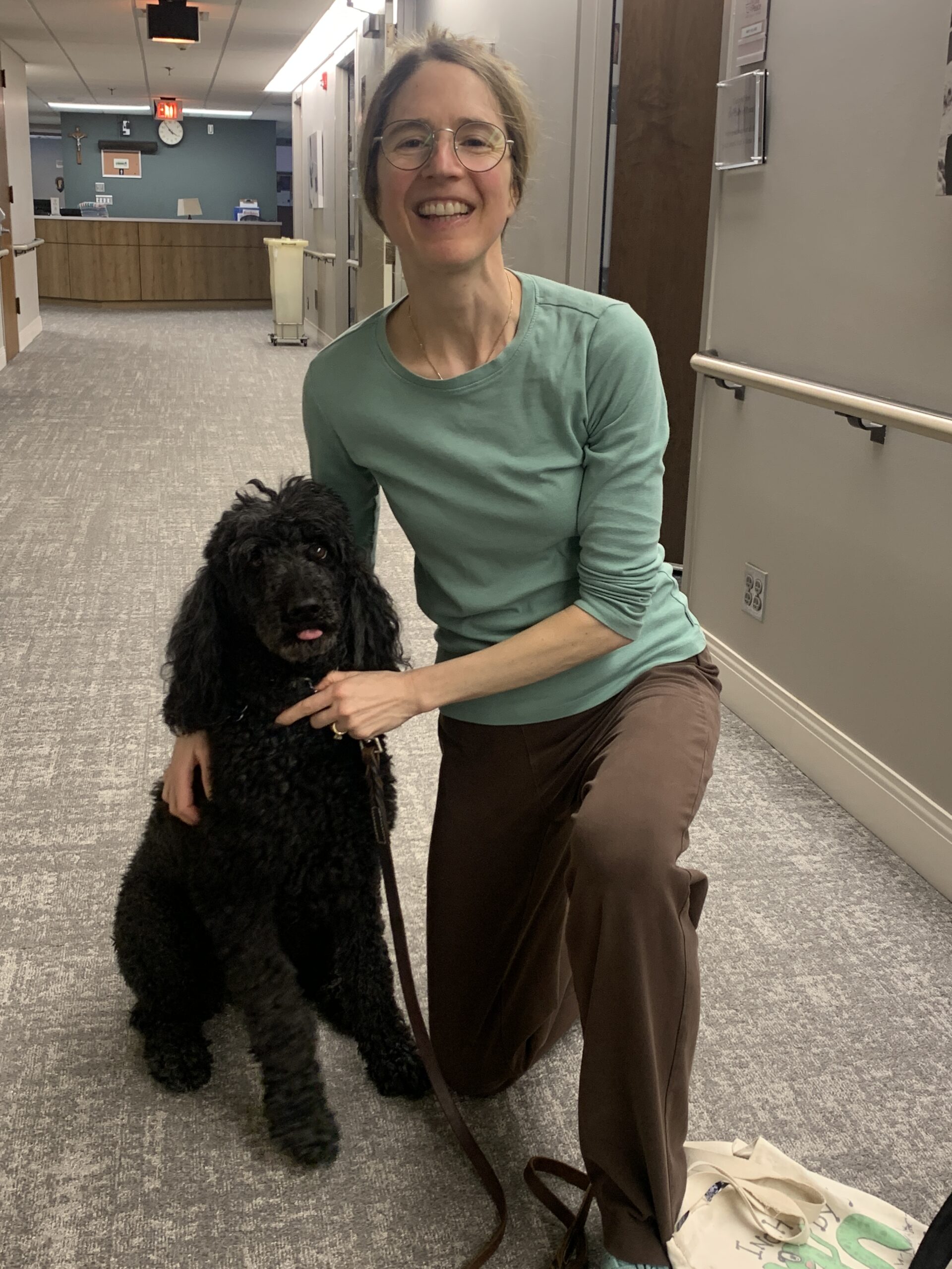Woman kneeling beside a medium black poodle with her tongue peaking out