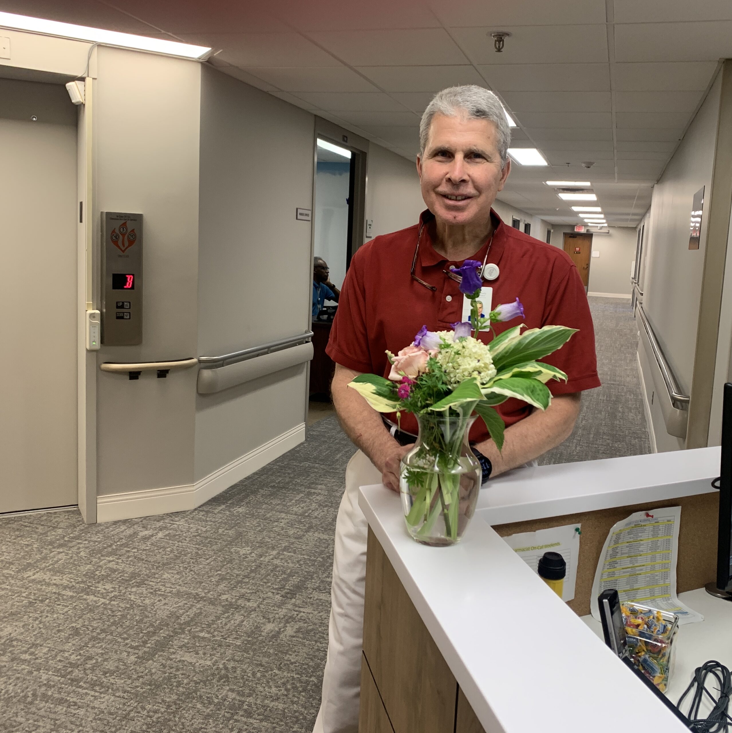 Picture of a male nurse wearing burgundy shirt and white pants by the nurses station with a plant