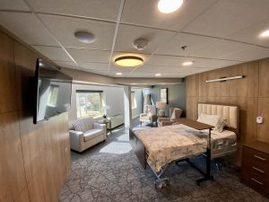 A picture of one of our rooms in the residence. It is a room full of sunlight with a bed, a TV on the wall, and chairs to the otherside of the bed along with two windows.