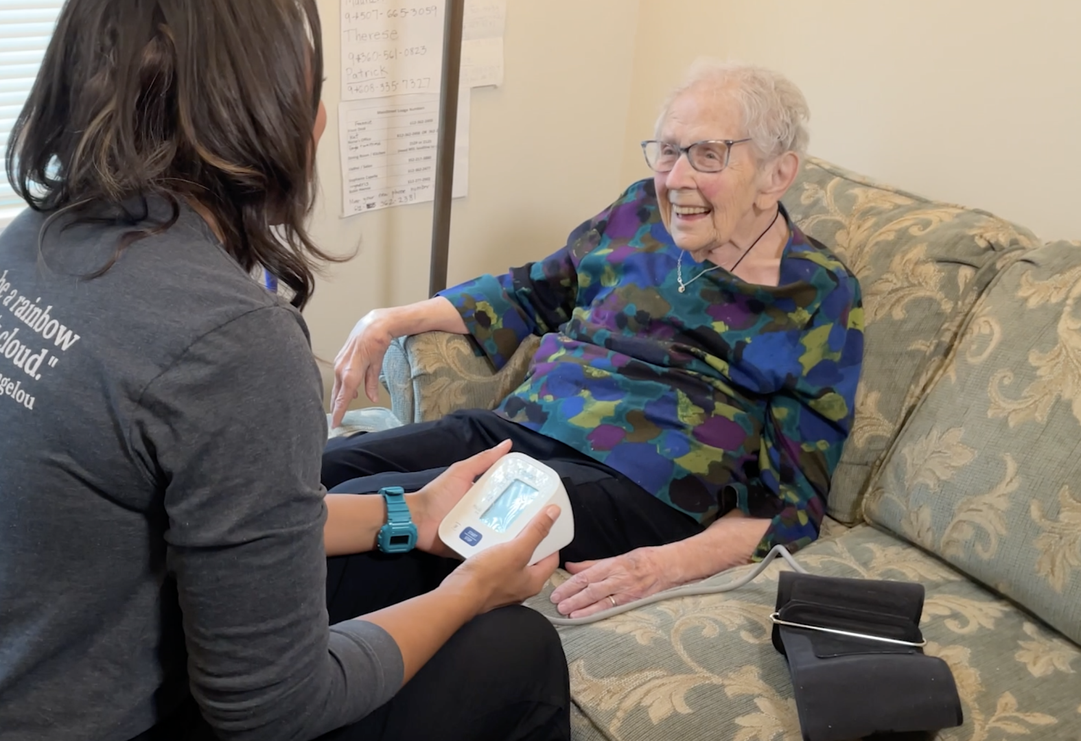 An elderly woman smiling at a caregiver. She is wearing glasses, blue and black blouse with black pants. Sitting on her couch. She is getting ready to have her blood pressure taken