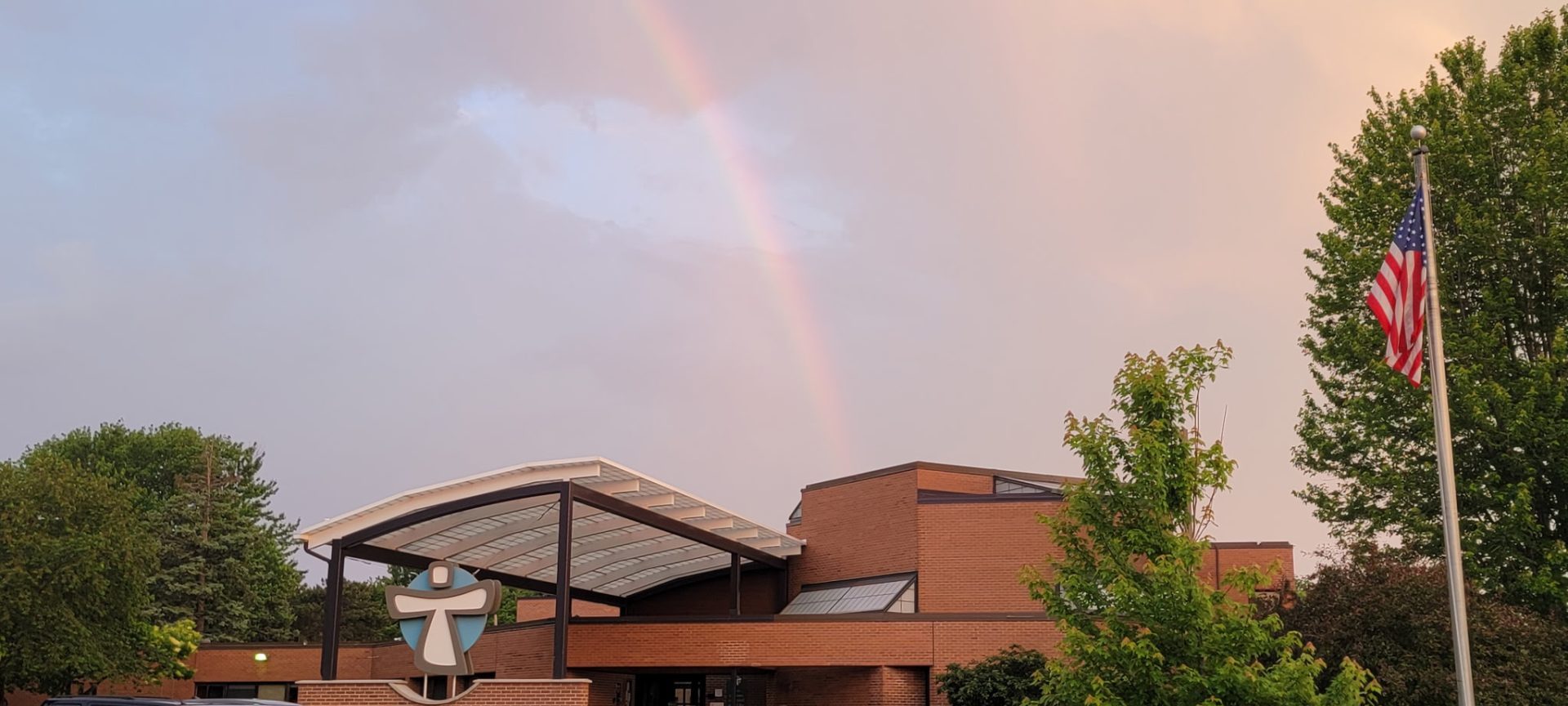 Picture of a red brick building with a rainbow coming down on top of it. Building is surrounded by green full trees
