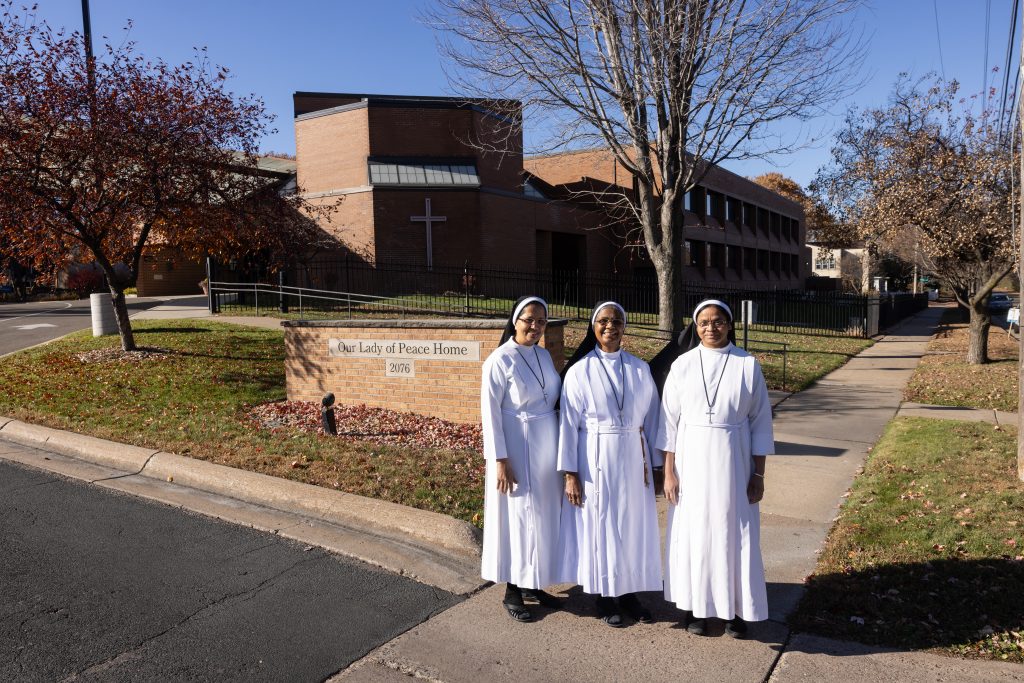 Four nuns in white habits stand together outside Our Lady of Peace Home in front of a brick building with a sign nearby